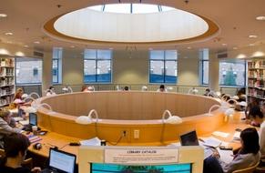 Students studying at large round wood counter, individual lamps at each work spot. Large round skylight above, windows along the back, book stacks on each side