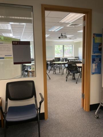 Looking through a door into a classroom full of round tables with chairs around them, windows looking into red square, projector on the ceiling, 222 on the door