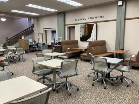 Room with tables, chairs, upholstered chairs and ottomans, booths, colorful art on the walls, "Learning Commons" on the wall and stairs in the background