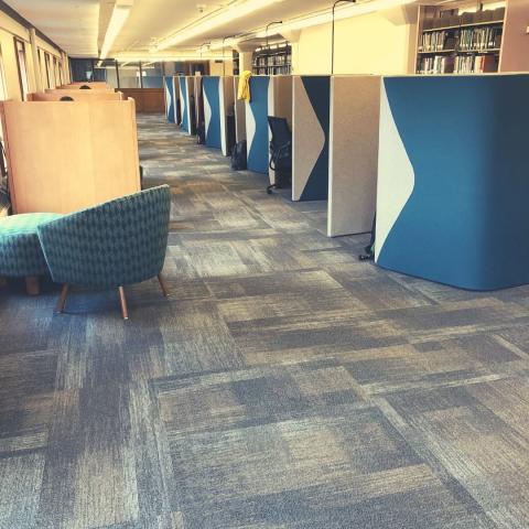 Row of individual wood carrels next to windows, study pods across an isle, group study room at the back, upholstered chair and ottoman in front