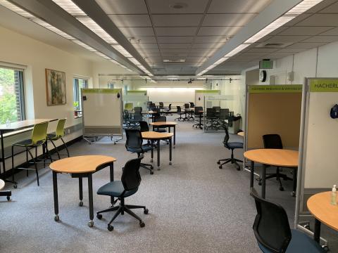 An open space with windows on left wall. Circular tables with chairs and whiteboard throughout. Entry into HH 210a at back of room.