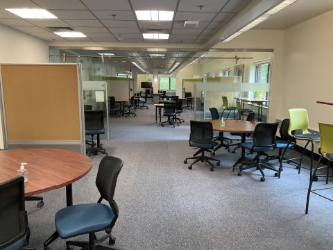 An open space with round tables surrounded by chairs scattered around. Back of whiteboard on far left. Glass sliding door divides the space.