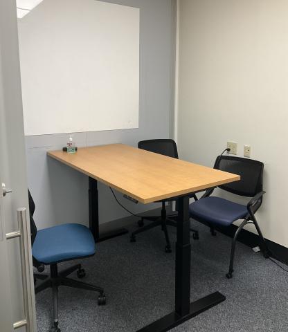 A small space with a tall table surrounded by chairs. A whiteboard is on the back wall above the table.