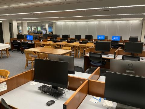 An open space with rows of computer desks in the foreground and back of the space. Long tables surrounded by chairs in middle of space.