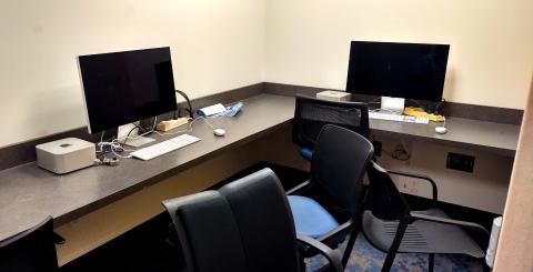 A small room with an L-shaped desk along the wall, two computer, two chairs. Mac video editing tools are on the desktop.