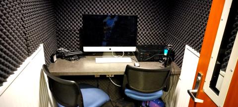 A small dark room with soundproof padding on the walls. A desk with a Mac, two chairs, a microphone fills most of the space.