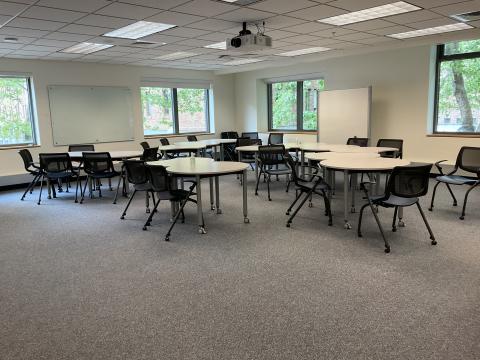 A classroom with crescent-shaped tables and chairs pushed together in corner of room. Whiteboard on right and left walls. Windows evenly spaced along walls..
