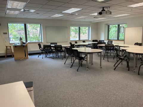 A classroom with crescent-shaped tables and chairs pushed together in corner of room. Teaching podium on back left. Whiteboard on back and right walls.