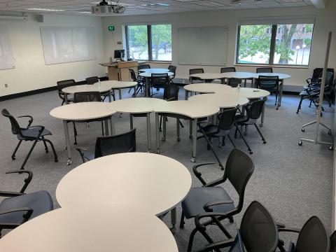 A classroom with crescent-shaped tables and chairs pushed together. Teaching podium in background. Two whiteboards on left wall, one on back wall, one on right.