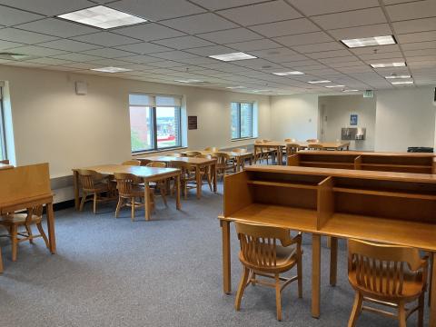 An open space with carrels in the right foreground and long tables along lefthand wall, each with four chairs. Water fountains and bathrooms on back wall.