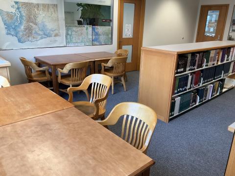 A small corner space with two long tables with chairs, each against a wall. Short bookstack in center of space. Maps on wall, doors to offices on left.