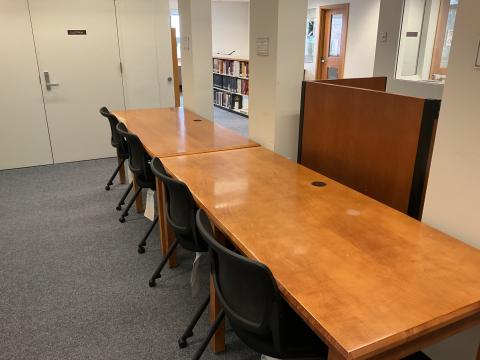 A very long table, with four chairs on one side, pressed up against columns in a narrow space. Bookstack and office in background.
