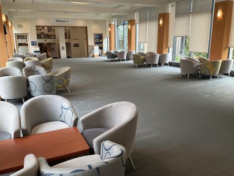 A wide-open space. Many floor-to-ceiling windows. Coffee tables surrounded by lounge chairs spaced along walls. Entrance to Wilson Library in back.