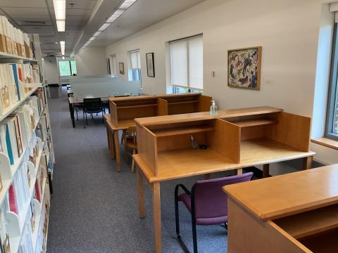 Three rows of carrels lined against right wall in foreground. Rows of long tables with frosted glass dividers in distance. Bookstack on left. 