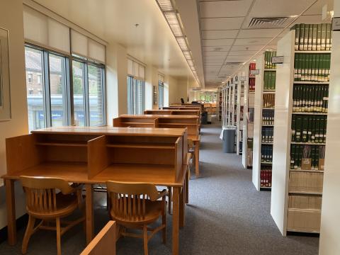 An open space with lots of natural light. Windows and carrels line the left wall with book stacks on the right, making a narrow walkway.