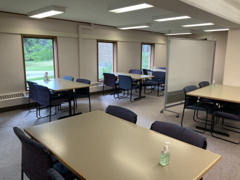 Tables surrounded by four chairs against windowed wall on right and in foreground. Whiteboard on right.
