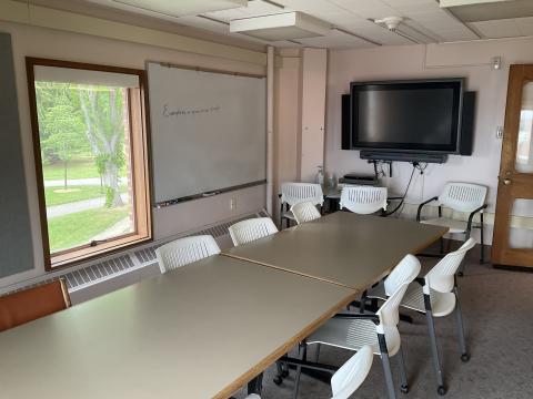 A long table surrounded by chairs. There's a Smartboard on the opposite wall, a whiteboard and window on the left.