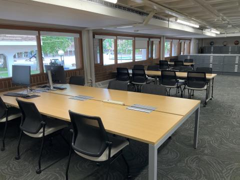 Long tables protruding from left wall, surrounded by chairs. Nearest table has two computer monitors on the far end. File cabinets in background.