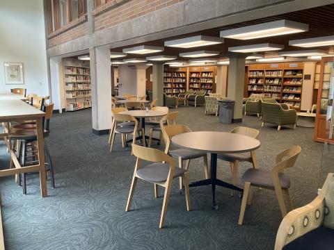 An open space. Round tables with chairs in middle of room. Counter with chairs on left against window. Armchairs with coffee tables in back near bookshelves.