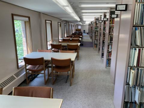 A narrow walkway with windows and tables extending from left wall, each with four chairs. Book stacks line the right side.