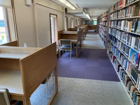 A narrow walkway with windows and carrels extending from left wall. Book stacks line the right side.