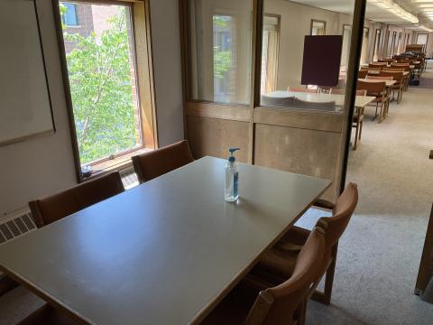 A small space with a long table surrounded by chairs in the middle. Whiteboard and window on left wall. Window and door into HH 270 on far side.
