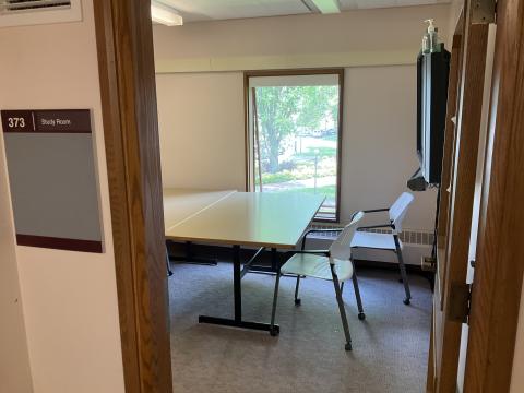 A small space, viewed from doorway, with two tables pushed together, surrounded by chairs. Smartboard on righthand wall, window on back wall; 373 Study Room