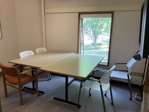 Small space with two tables pushed together, surrounded by chairs. Smartboard on right wall, window on back wall. Whiteboard on left wall.