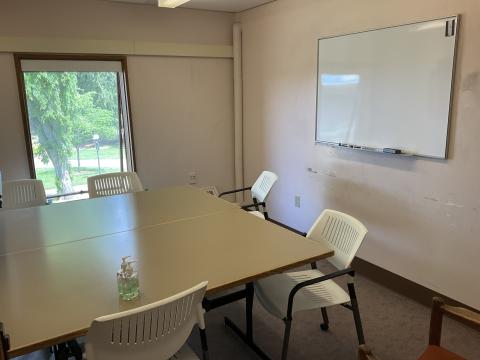 A small space with two tables pushed together, surrounded by chairs. Whiteboard on right wall, single window on back wall. 