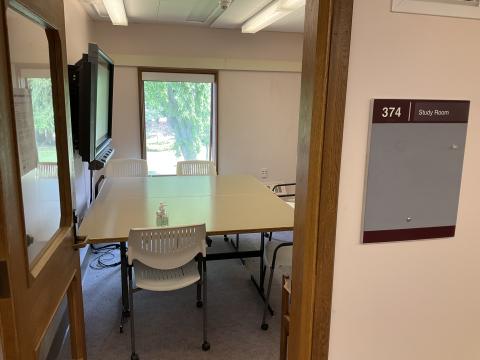 A small space, viewed from doorway, with two tables pushed together, surrounded by chairs. Smartboard on left wall, single window on back wall. 