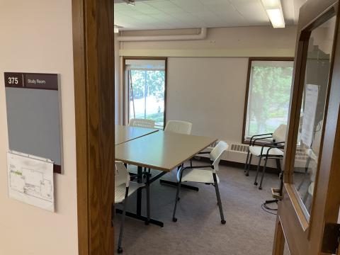 A small study room from the outside with two desks pushed together, chairs surrounding. Two small windows in back wall; 375 Study Room.