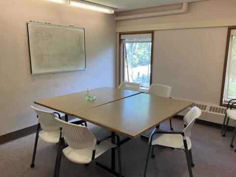 A small study space with two tables pushed together in the middle, surrounded by chairs. Whiteboard on left wall. small windows on back wall.