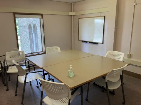 A small space with two tables pushed together, surrounded by chairs. Whiteboard on righthand wall, single window on back wall. 