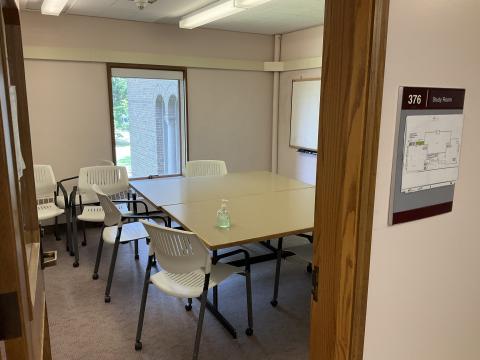 Small space from outside doorway with two tables pushed together, surrounded by chairs. Whiteboard on right wall, single window on back wall 