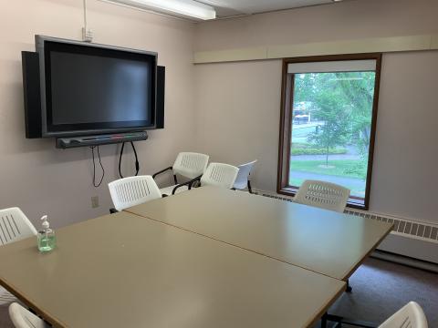 Small space with two tables pushed together, surrounded by chairs. Smartboard on left wall, window on back wall.