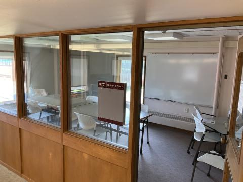 A windowed room with a whiteboard on the back wall, a long table with multiple chairs in the middle, windows looking outside on back and left wall.