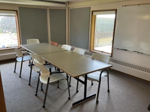 A room with a long conference table surrounded by multiple chairs. Small window, felt board on the left wall. Whiteboard, felt board, small window on back wall.