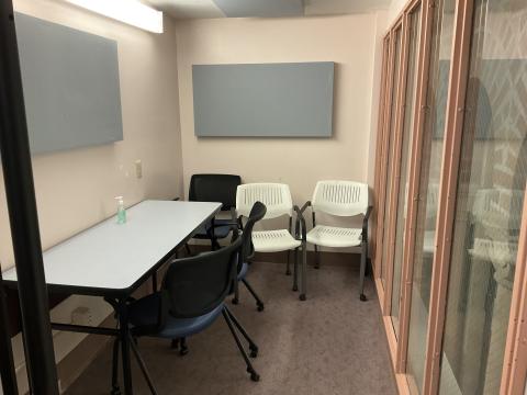 Very small space with table against left wall, five chairs scattered in the space, a cloth rectangles on the left and back wall. Right wall is a window.