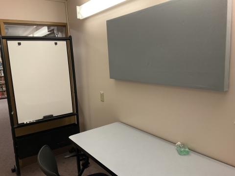 A very small space with a long table against the wall, a felt rectangular wall-hanging on wall above table. Whiteboard on left.