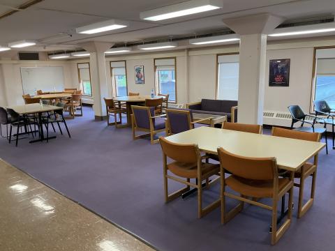 Long open space with rectangular tables and chairs. Round tables on back-left. Windows on back wall. Columns throughout the space. Carpeted floor.