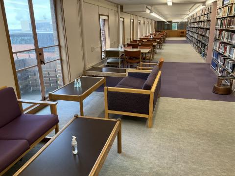 Long narrow space, large windows on left wall. Couches and coffee tables in foreground, long tables on back left wall. Book stacks on the right.