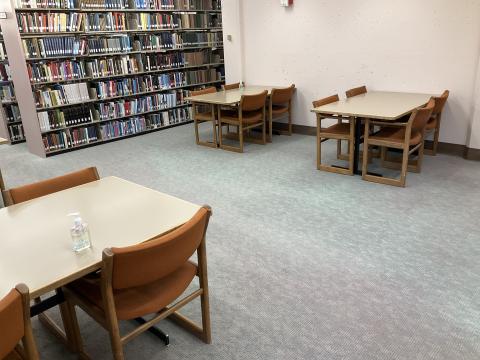 Open space with three tables, two against back wall, one in foreground. Bookstacks on the back-right.