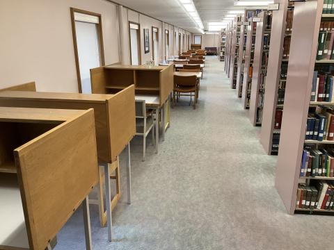 A narrow space with carrols on front left, long tables along remaining left wall. Bookstacks on the right. Walkway between tables and bookstacks.
