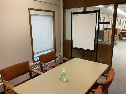 A small room with long table surrounded by chairs. Open doorway on right. Whiteboard against window into room. Window outside on the left.