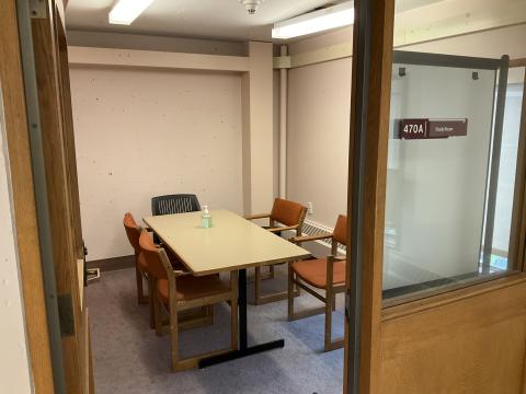 A small room viewed from outside the doorway. Window into the room on the right. Long table surrounded by chairs. Whiteboard blocking window into room.