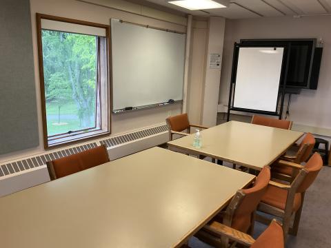 Two tables pushed together to form long table, surrounded by chairs. Large whiteboard on left wall, small one on back wall. Window on left wall.