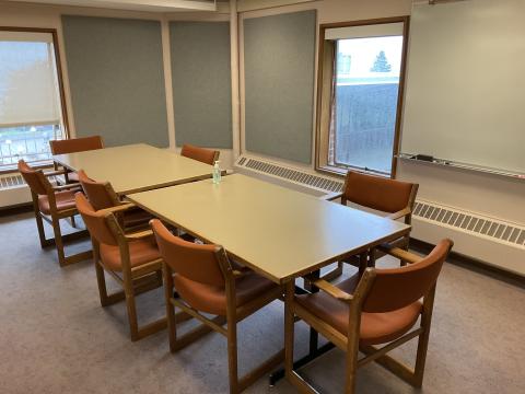 Two tables pushed together to form long table, surrounded by chairs. Windows on back and right wall. Felt padding on back and right wall.