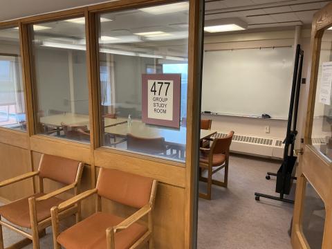 A room viewed through open doorway. Windowed wall into room on left side of door. Long table surrounded by chairs. Whiteboard on back wall.