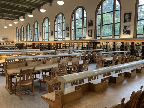 A very large room with many long, slanted reading desks with lamps. Large arched windows and bookshelves along back wall. 