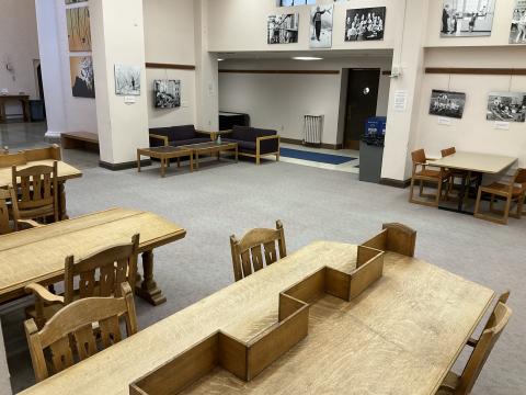 An open space with sparse furniture. Long, slanted reading desks the foreground. Standard long table on back right. Couch on back left. Hallway in background.